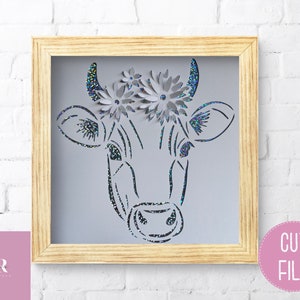 May include: A black and white line drawing of a cow's head with a floral crown. The cow's head is framed in a light wood frame. The image is a cut file.