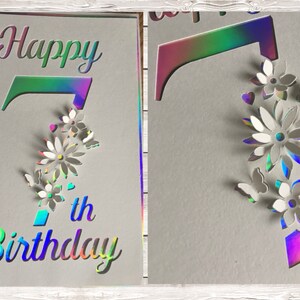 Op de afbeelding: Een holografische kaart met de woorden "Happy 7th Birthday" in een regenboogkleurig lettertype. Het cijfer 7 is uitgesneden en versierd met witte papieren bloemen en vlinders.
