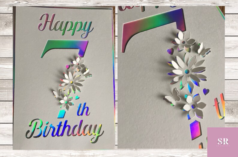Op de afbeelding: Een holografische kaart met de woorden "Happy 7th Birthday" in een regenboogkleurig lettertype. Het cijfer 7 is uitgesneden en versierd met witte papieren bloemen en vlinders.