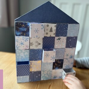 May include: A blue and white paper house shaped advent calendar with 24 numbered doors. Each door has a different winter-themed pattern. The calendar is on a wooden surface. The text "CUT FILE" and "SVG" are visible in pink circles.