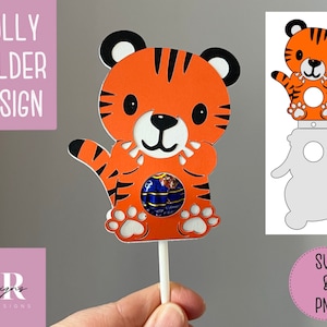 Puede incluir: Diseño de soporte para piruletas de tigre naranja y negro con un hueco para un caramelo. El diseño incluye una cara de tigre con una expresión linda y huellas de patas. El soporte está en un palo blanco. La imagen también muestra una plantilla separada para el soporte.