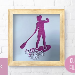 May include: A pink silhouette of a person paddleboarding with flowers around the board. The image is in a light wood frame.