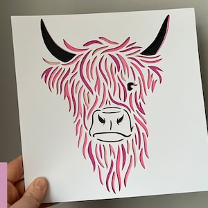 Puede incluir: Un dise&ntilde;o de corte de papel rosa y negro de la cabeza de una vaca de las tierras altas. El dise&ntilde;o es intrincado y detallado, con el pelo de la vaca representado por papel en capas.