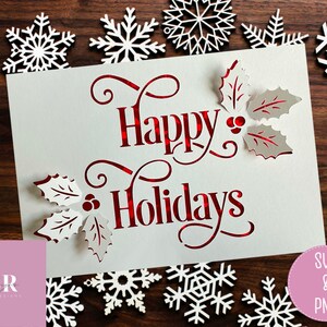 Puede incluir: Una tarjeta blanca con letras rojas que dice "Happy Holidays" con hojas de acebo y bayas recortadas alrededor del texto. La tarjeta est&aacute; rodeada de copos de nieve blancos sobre un fondo de madera.