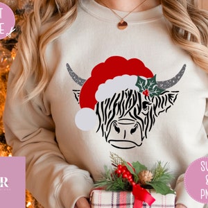 K&ouml;nnte beinhalten: Ein wei&szlig;er Pullover mit einer schwarz-wei&szlig;en Highland-Kuh, die einen roten Weihnachtsmann-Hut mit einem gr&uuml;nen Stechpalmenzweig tr&auml;gt. Die Kuh schaut nach rechts. Der Pullover wird von einer Person mit langen blonden Haaren getragen.