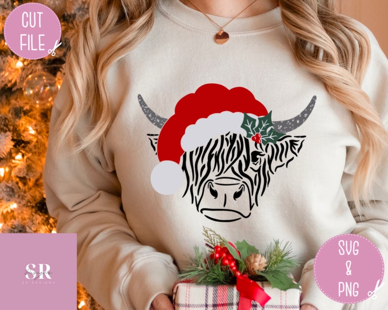 K&ouml;nnte beinhalten: Ein wei&szlig;er Pullover mit einer schwarz-wei&szlig;en Highland-Kuh, die einen roten Weihnachtsmann-Hut mit einem gr&uuml;nen Stechpalmenzweig tr&auml;gt. Die Kuh schaut nach rechts. Der Pullover wird von einer Person mit langen blonden Haaren getragen.