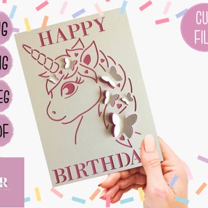 May include: A pink and white card with the text "Happy Birthday" and a cut-out design of a unicorn with butterflies. The card is being held by a hand.