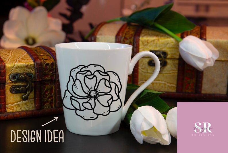 May include: White ceramic mug with a black outline of a flower design. The mug is sitting on a table with a wooden box and white tulips in the background. The text "DESIGN IDEA" is visible in the lower left corner of the image.