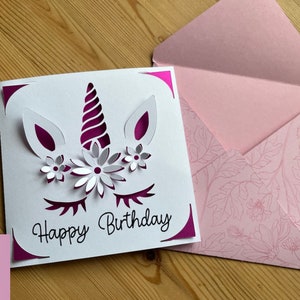 May include: A white card with a pink unicorn design and the text "Happy Birthday". The card is folded in half and has a pink envelope with a floral pattern.