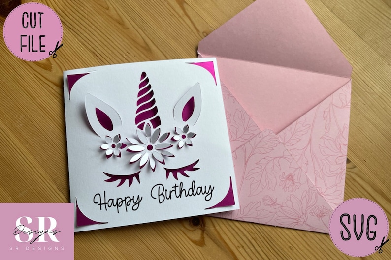 May include: A white card with a pink unicorn design and the text "Happy Birthday". The card is folded in half and has a pink envelope with a floral pattern.