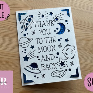 Op de afbeelding: Een witte kaart met zwarte inkt en blauwe glitteraccenten. De kaart heeft een raket, sterren, planeten en een halve maan. De tekst op de kaart luidt "Thank You To The Moon And Back".