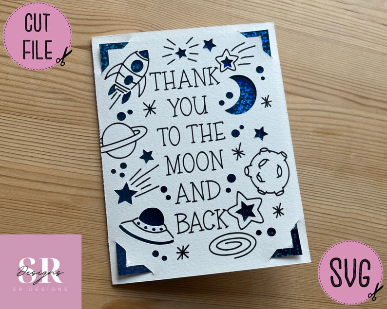 Op de afbeelding: Een witte kaart met zwarte inkt en blauwe glitteraccenten. De kaart heeft een raket, sterren, planeten en een halve maan. De tekst op de kaart luidt "Thank You To The Moon And Back".