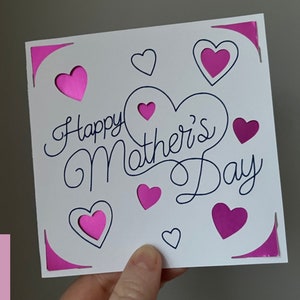May include: A white card with the text "Happy Mother's Day" in black cursive script. The card is decorated with pink hearts, some of which have a cut-out design.