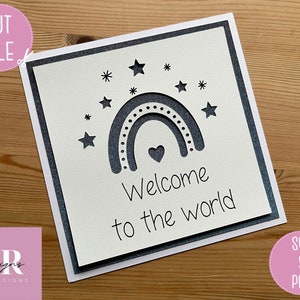 May include: A white card with a gray border and a cut-out design of a rainbow with stars and a heart. The text "Welcome to the world" is written below the rainbow.