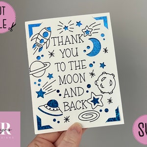 Op de afbeelding: Een witte kaart met een blauwe glitterrand en zwarte tekst die "Bedankt tot aan de maan en terug" luidt. De kaart is versierd met sterren, een raket, een planeet, een maan en een UFO.