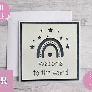 May include: A white card with a black and white rainbow, stars, and a heart. The text "Welcome to the world" is written below the rainbow.