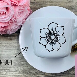 May include: White ceramic teacup and saucer with a black floral mandala design. The design is centered on the cup and features a circular pattern with intricate details. The cup and saucer are on a wooden surface.