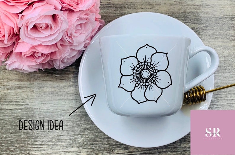 May include: White ceramic teacup and saucer with a black floral mandala design. The design is centered on the cup and features a circular pattern with intricate details. The cup and saucer are on a wooden surface.