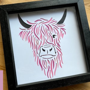 Puede incluir: Una silueta de cabeza de vaca Highland rosa y blanca recortada en papel y enmarcada en un marco negro.