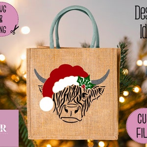 K&ouml;nnte beinhalten: Eine Jute-Tragetasche mit einem Design einer schottischen Hochlandkuh, die einen Weihnachtsmann-Hut mit Stechpalme tr&auml;gt. Die Tasche hat einen gr&uuml;nen Griff.