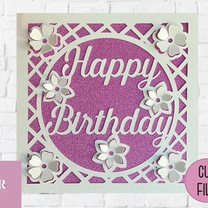 Op de afbeelding: Witte papieren uitsnijkaart met een paarse glitterachtergrond. De kaart heeft een cirkel met de woorden "Happy Birthday" in wit handschrift. De cirkel is omgeven door een wit tralieontwerp en witte papieren bloemen.