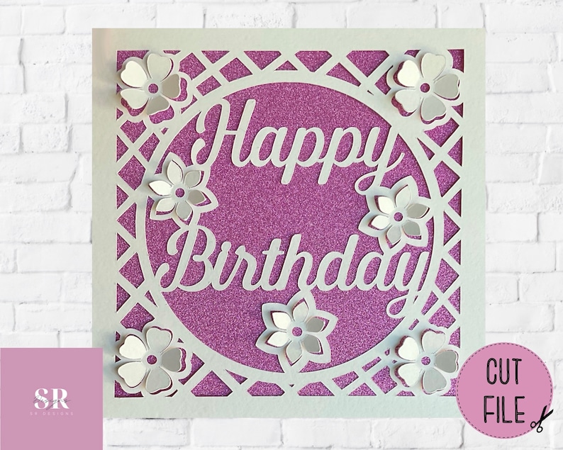Op de afbeelding: Witte papieren uitsnijkaart met een paarse glitterachtergrond. De kaart heeft een cirkel met de woorden "Happy Birthday" in wit handschrift. De cirkel is omgeven door een wit tralieontwerp en witte papieren bloemen.