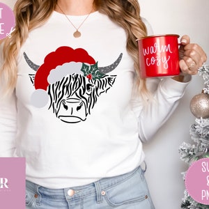 K&ouml;nnte beinhalten: Ein wei&szlig;es Langarmshirt mit einer Schwarzwei&szlig;illustration einer schottischen Hochlandkuh, die einen roten Weihnachtsmann-Hut mit einem gr&uuml;nen Stechpalmenzweig tr&auml;gt. Der Text "warm cozy" befindet sich auf einer roten Tasse.