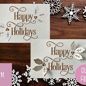 Puede incluir: Dos tarjetas blancas con el texto "Happy Holidays" en letras marrones y un dise&ntilde;o de hoja de acebo. Las tarjetas est&aacute;n sobre una superficie de madera marr&oacute;n con copos de nieve blancos esparcidos a su alrededor. El texto "CUT FILE" est&aacute; en un c&iacute;rculo rosa en la esquina inferior derecha.