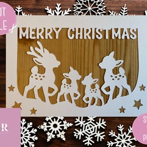 May include: White paper cut-out design featuring a family of deer with the words "Merry Christmas" at the top. The design is set against a woodgrain background.