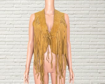 May include: Mustard-colored suede vest with long fringe detailing. The vest has a V-neck and a tie closure at the front. The fringe hangs vertically, adding a bohemian flair to the garment. The vest is displayed on a mannequin.