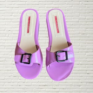 May include: A pair of lavender patent leather sandals with a black buckle. The sandals have a C. Label logo on the strap.