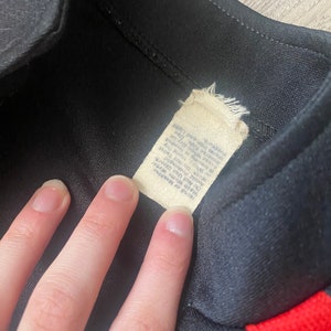 K&ouml;nnte beinhalten: Schwarze und rote Trainingsjacke mit einem wei&szlig;en Etikett mit der Aufschrift "Made in Korea, 100% Polyester, Maschinenw&auml;sche kalt, Trocknergeeignet bei niedriger Temperatur, Nicht bleichen, Nicht b&uuml;geln".