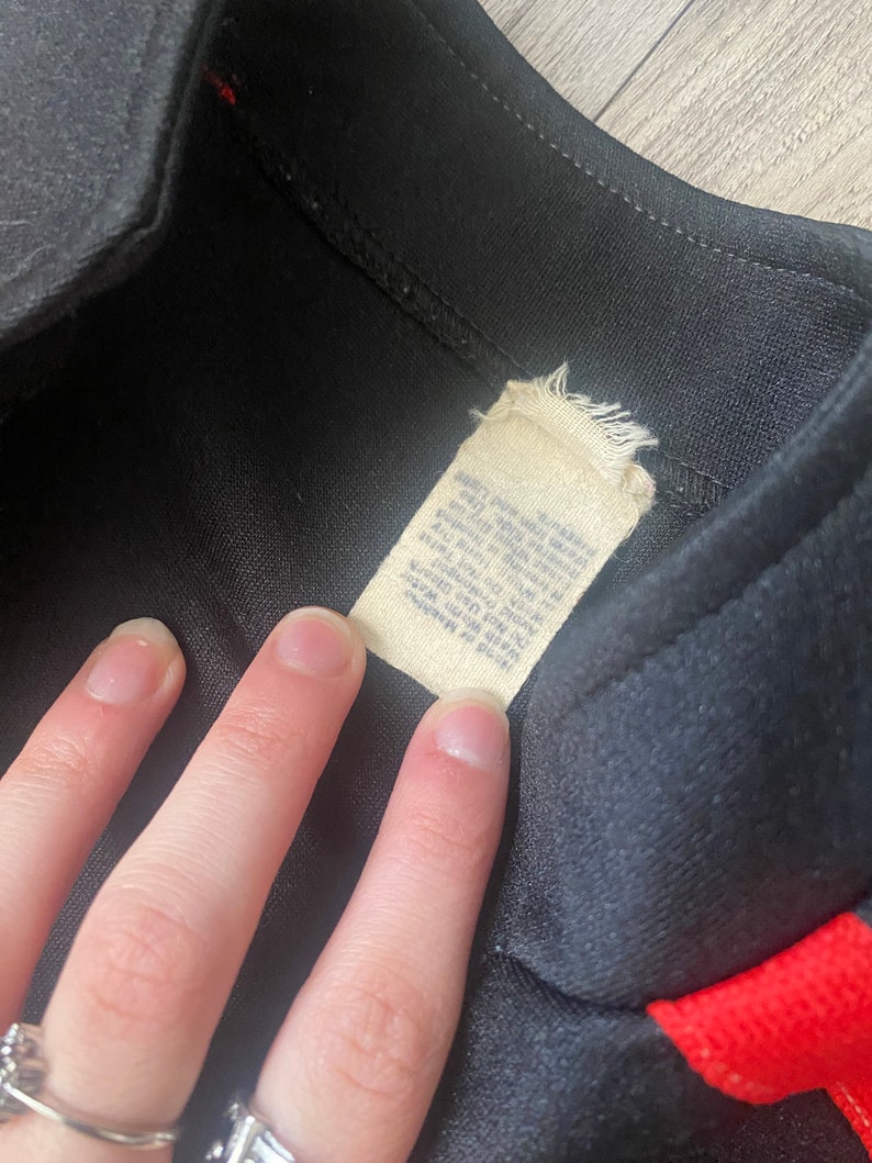 K&ouml;nnte beinhalten: Schwarze und rote Trainingsjacke mit einem wei&szlig;en Etikett mit der Aufschrift "Made in Korea, 100% Polyester, Maschinenw&auml;sche kalt, Trocknergeeignet bei niedriger Temperatur, Nicht bleichen, Nicht b&uuml;geln".