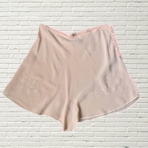 Könnte beinhalten: Hellrosa Shorts mit zarter Blumenstickerei. Die Shorts haben einen weichen, fließenden Stoff und eine bequeme Passform. Die Stickerei ist weiß und verleiht dem Kleidungsstück einen Hauch von Eleganz.