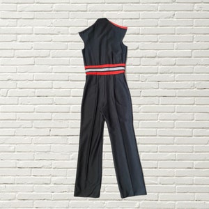 K&ouml;nnte beinhalten: Schwarzer Jumpsuit mit einem roten und wei&szlig;en Streifen um die Taille. Der Jumpsuit hat einen hohen Kragen und kurze &Auml;rmel.