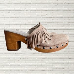 May include: Beige suede clogs with a wooden platform and fringe detail. The clogs have a high heel and are decorated with metal studs.