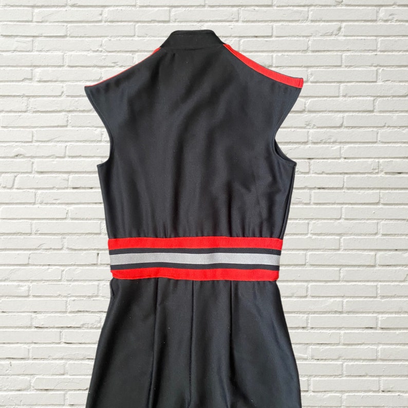 K&ouml;nnte beinhalten: Schwarzer &auml;rmelloser Jumpsuit mit einem roten und silbernen Streifen um die Taille.