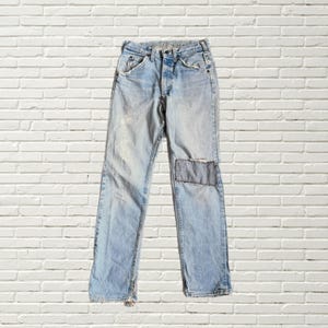 May include: Light blue denim jeans with a faded wash and a patched knee. The jeans have a button fly, classic five-pocket design, and distressed detailing at the hem. The jeans are displayed against a white brick wall.