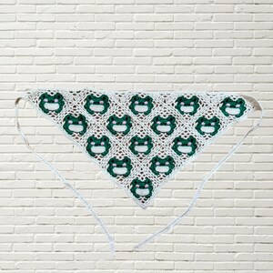 May include: A white crochet bandana with a green frog pattern. The frog faces have white eyes and pink mouths. The bandana has two long, light blue ties.