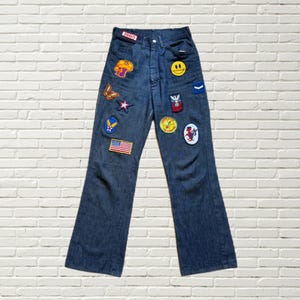 May include: Dark wash denim jeans with various patches, including a mushroom, butterfly, star, smiley face, and an American flag. The jeans have a high waist and a flared leg. The year "1980" is printed on the jeans.