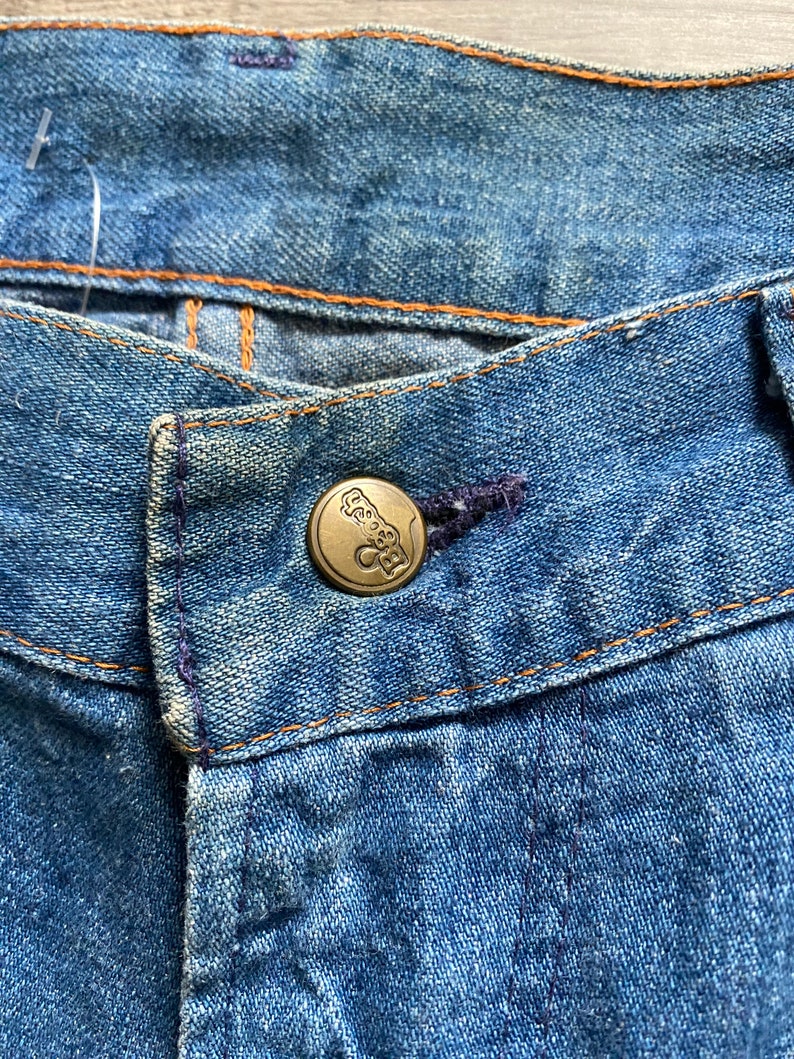 May include: Close-up of a pair of blue denim jeans with a metal button featuring a logo. The button is located at the top of the zipper fly.