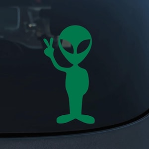 May include: Green alien sticker with a peace sign, on a dark grey car window.