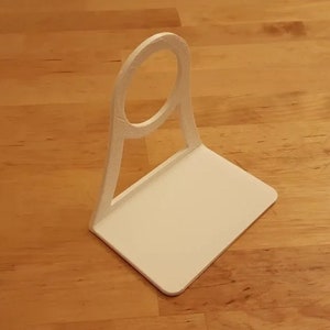 May include: White plastic phone holder with a circular cutout for a phone. The holder has a flat base and a curved back.