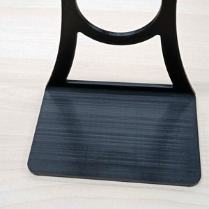 May include: A black plastic phone stand with a circular cutout for holding a phone. The stand has a flat base and a curved back.