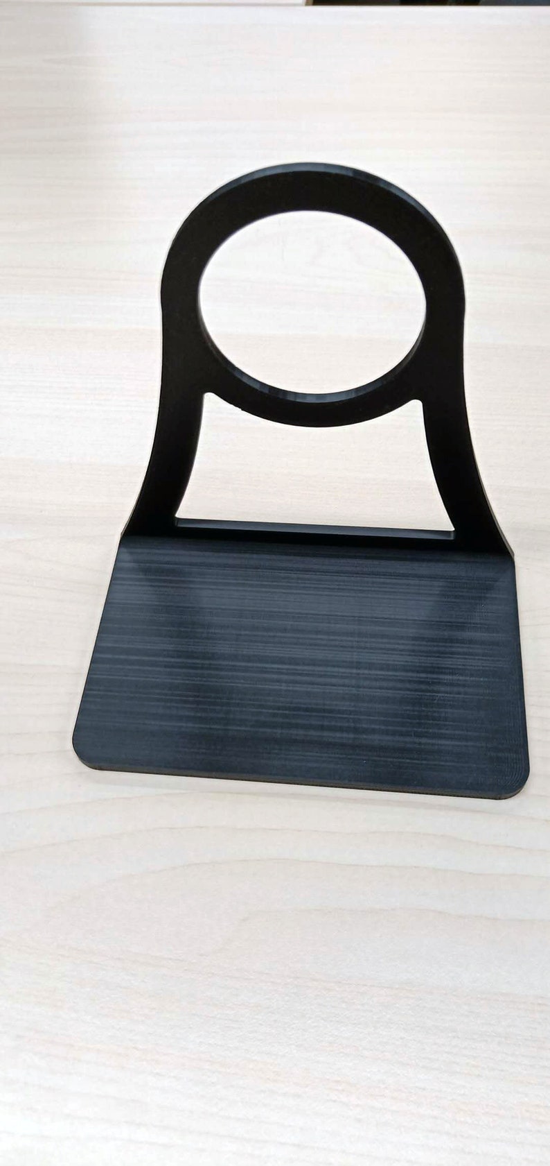 May include: A black plastic phone stand with a circular cutout for holding a phone. The stand has a flat base and a curved back.