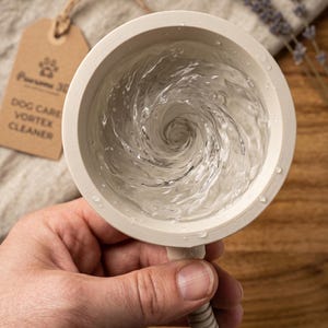 Puede incluir: Un limpiador vortex circular de color beige para perros con un remolino de agua en su interior. Una etiqueta de papel marrón unida al dispositivo presenta un icono de huella y el texto: Pawsome 3D, DOG CARE VORTEX CLEANER. Tubo acanalado.