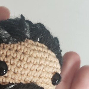 Puede incluir: Una figura amigurumi de crochet de una persona con cabello negro, una larga barba negra con vetas blancas y ojos negros. La figura lleva una camisa beige.
