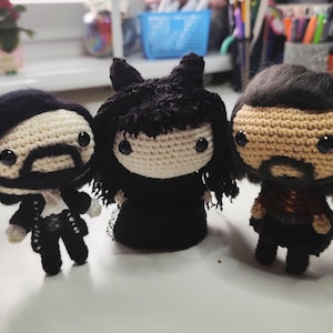 Puede incluir: Tres juguetes de peluche de crochet, cada uno con cabello negro y una expresión facial diferente. El juguete de la izquierda tiene un bigote y una barba negros, el juguete del medio tiene un vestido negro y el juguete de la derecha tiene una camisa roja.