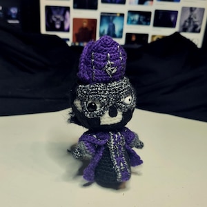 Può includere: Un giocattolo di peluche all'uncinetto con un mantello con cappuccio viola e nero, una maschera argentata e un cappello viola. Il giocattolo ha un viso bianco con occhi neri e una bocca nera.