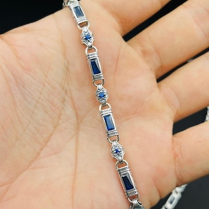 Può includere: Un braccialetto d'argento con pietre preziose blu rettangolari e rotonde. Il braccialetto è tenuto in una mano su sfondo nero. Il design del braccialetto presenta pietre blu rettangolari e rotonde alternate.